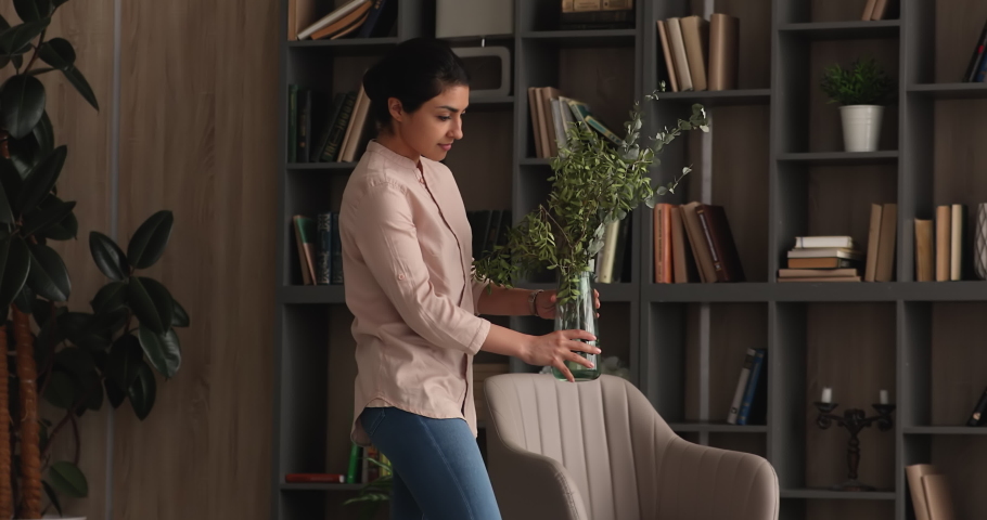 Smiling tidy young indian ethnicity female homeowner putting vase with greens and flowers on table in modern living room, making interior more comfortable, decorating styling improving accommodation. Royalty-Free Stock Footage #1068159926