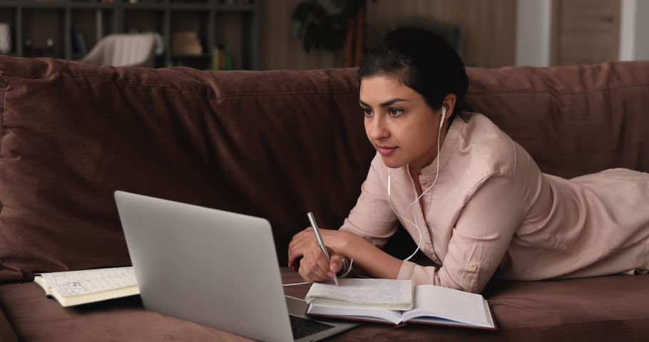 Concentrated millennial indian woman in wired earphones lying on sofa with books and laptop, involved in watching educational online lecture, improving knowledge distantly, e-learning concept. | Shutterstock HD Video #1068159977
