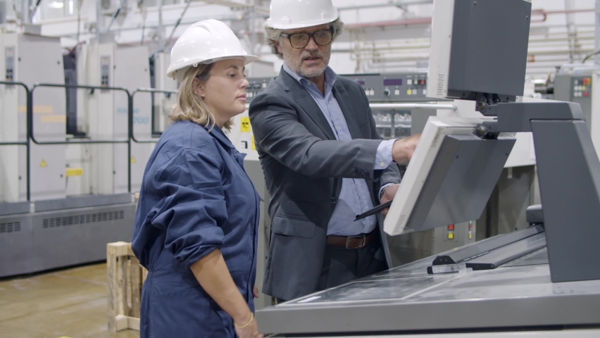 Older engineer teaching factory employee to operate new machine on plant floor, showing guide on tablet screen, using control panel. Production process or machinery concept Royalty-Free Stock Footage #1068161198