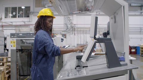Confident black female factory worker operating industrial machine, working at control board, using tablet. Production process or machinery concept