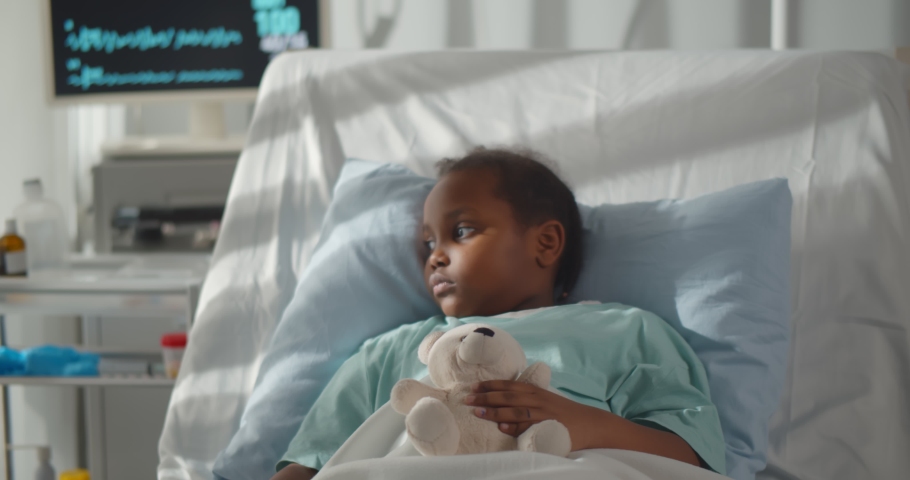Portrait of sick african girl hugging teddy bear lying in hospital bed. Ill and sad afro-american child patient resting in intensive care unit at modern pediatrics department Royalty-Free Stock Footage #1068162743