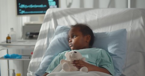 Portrait of sick african girl hugging teddy bear lying in hospital bed. Ill and sad afro-american child patient resting in intensive care unit at modern pediatrics department