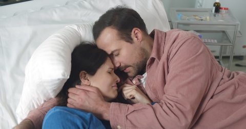 Husband hugging and comforting crying sick woman lying together in hospital bed. Man visiting ill wife patient feeling scared and depressed resting in ward