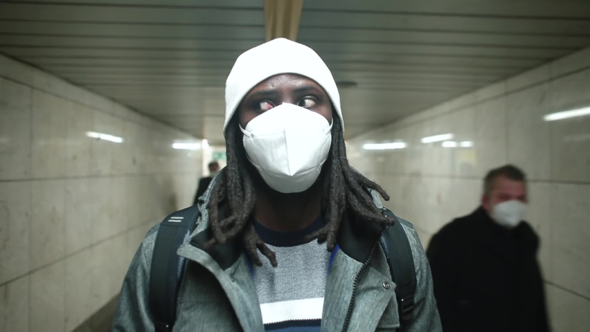 Black man face mask moves through tunnel in city underground. People walking nearby. Moving passengers in passage. Move between levels. Change of the train line. Camera movement.