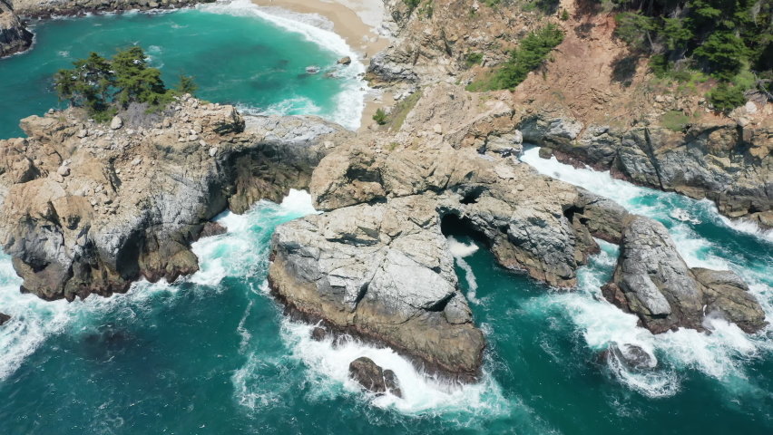 Ocean cliffs surrounded by beautiful deep blue waters. Sea waves crashing with splashes on rocky shore with secluded small sandy beach under the bluff. Stormy beach and cliffs aerials, Pacific ocean