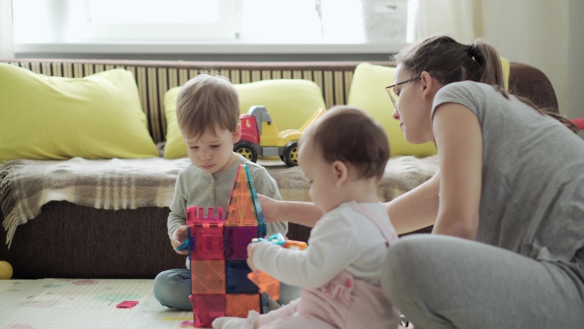 Childhood, family, motherhood, parenthood concept - young happy mother enjoy playing magnets blocks builds castle with toddler kids. Three Children have fun together in playroom floor on quarantine Royalty-Free Stock Footage #1068168230