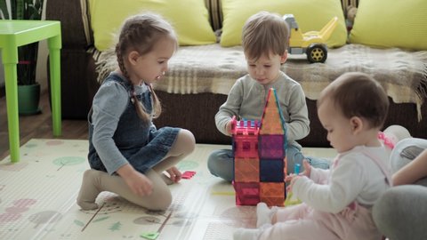 Childhood, family, motherhood, parenthood concept - young happy mother enjoy playing magnets blocks builds castle with toddler kids. Three Children have fun together in playroom floor on quarantine