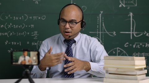 Indian young teacher man sitting wearing headset teaching online video conference live stream by smartphone. Asian teacher teaching mathematics class webinar online for students learning.