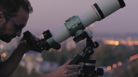 Astronomer with a telescope watching at the stars and Moon with blurred city lights in the background.	
