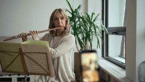 Woman playing flute and staying in apartment room during covid pandemic. American middle aged female using musical instrument and looking at book while sitting in front of smartphone screen in light