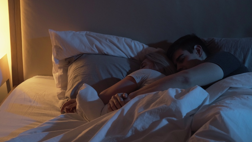 Sleeping couple. Happy marriage. Tender relationship. Relaxed man enjoying to embrace cuddle tired wife lying in cozy bed at night in dark bedroom with blue warm yellow lamp light. | Shutterstock HD Video #1068179573
