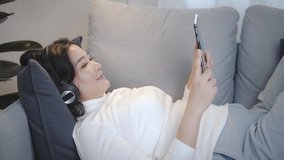 Happy cheerful Asian woman wearing wireless headphones watching to the movie from a tablet in relax time at home lifestyle and pleasure concept