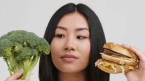 Unhealthy Food Choice. Hungry Asian Lady Choosing Burger Vs Healthy Broccoli Standing Posing On White Studio Background. Chinese Lady Licking Lips Looking At Hamburger. Nutrition Concept
