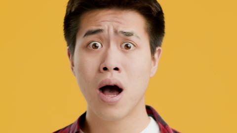 Portrait Of Shocked Asian Man Posing Looking Aside And At Camera In Shock Standing On Yellow Studio Background. Closeup Of Chinese Man With Embarrassed Emotional Face Concept