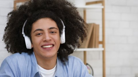 Bushy African American Guy Wearing Wireless Headphones Listening To Music And Smiling Looking At Camera Posing Sitting Indoors. E-Learning And Educational Online Podcast Concept