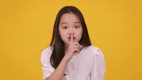Keep our secret. Cute little asian girl putting finger on lips, showing hush gesture at camera, orange studio background, slow motion