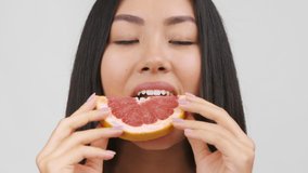 Asian Woman Eating Slice Of Juicy Grapefruit Enjoying Fresh Citrus Fruit Posing On White Studio Background, Smiling To Camera. Healthy Natural Nutrition, Organic Food And Vitamins Concept
