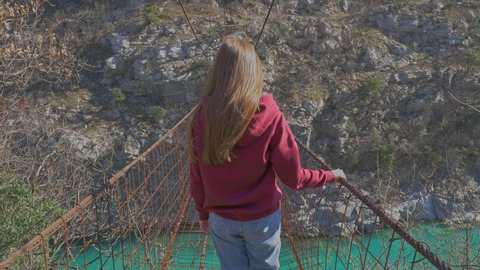 Young red hair woman walk through suspension bridge above the canyon in Montenegro in sunny day. Dangerous walking. Wild nature. Montenegro, Podgorica