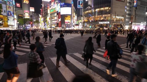 TOKYO - APRIL 03, 2018: Busy Shibuya scramble crossing at night time, walk through people crowd, overhead shot. Bright buildings of shopping district seen ahead. Diagonal crosswalk full of pedestrians