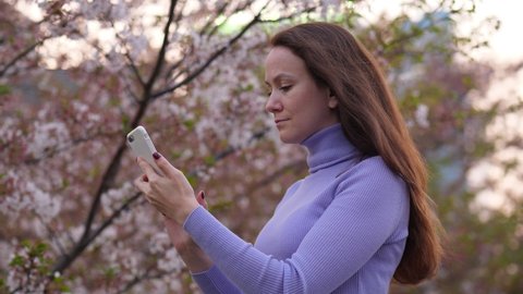 Pensive woman using phone, portrait shot against blossom cherry tree, blurred background. European lady stare to smartphone, stand at Tokyo alley. Beautiful hanami time at Japan in April