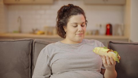 Untidy overweight woman eating hot dog at home, slovenly lifestyle, mess