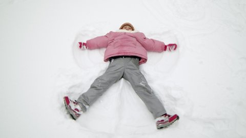 High angle full of seven-year-old Asian girl wearing winter clothes, lying on back on white snow outdoors, making snow angel, moving hands and legs to sides and back
