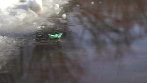 Melting snow and start of spring season concept. Big and huge puddles in snowy city park. Cute small green papper boat sailing in water on sidewalk