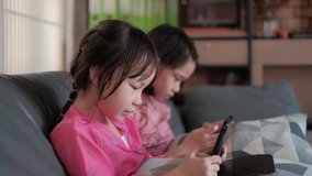 Two Asian girls in pink dresses are playing fun and exciting online mobile games at the gray sofa in their living room.