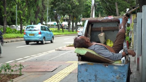 JAKARTA, INDONESIA - FEBRUARY 24, 2021:  A poor and old man works as a garbage scavenger is sleeping in his trash cart outdoor next to the public road.