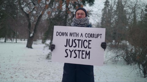 A Man is Standing in the Winter Park and Holding a Sign Down with a Corrupt Justice System. He is Protesting against the Government System of his Country. Standing against Corruption. Single Picket.