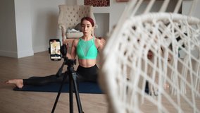 Young Slim Woman Blogger Conducts a Fitness Lesson Online, Taking Pictures on a Mobile Phone. Coach Blogger Leads a Stretching Workout at Home. Video Blogging Concept. Live Streaming. Social Distance.