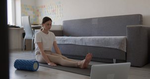 Attractive woman sitting on yoga mat and breathing deeply for relaxation after hard morning workout. Female athlete using wireless laptop during training at home.