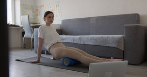 Attractive woman relaxing legs on massage roller after morning workout. Young brunette in activewear lying on yoga mat and watching video on wireless laptop.