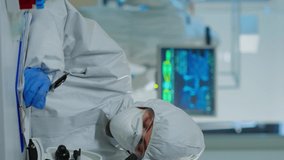 Vertical video: Medical research scientist conducting DNA experiments under microscope wearing protection suit and writing informations on clipboard. Chemist using high tech for vaccine development
