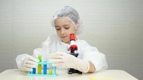 A child plays in a medical suit as a laboratory assistant. Kid conducts experiments by mixing multicolored chemicals in a flask and looking through a microscope. The concept of the development of