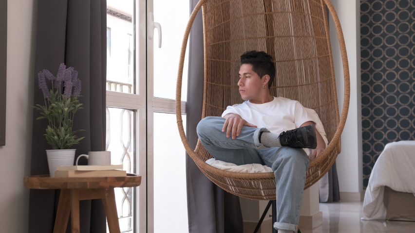 young man looking out the window sitting in a hanging chair at home