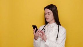 Female wearing coat stethoscope on shoulders looking at phone camera, isolated on yellow wall, doctor make video call interact through internet talk with patient provide help online counseling concept