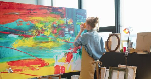Back view of Caucasian young pretty talented woman painter standing in own workshop drinking hot coffee or tea looking at finished abstract colorful painting. Contemporary art, art studio concept