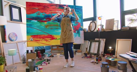 Rear of happy young Caucasian woman artist in apron dancing and rhythmically moving while listening to music in headphones and drawing picture on big canvas in own art studio, painter concept