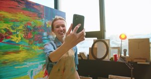 Close up of happy cheerful young beautiful talented Caucasian woman painter videochatting on smartphone talking on online video call in art studio after painting on big canvas, artist concept