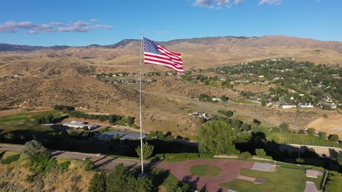 Drone camera gliding back from an American Flag waving on the top of a hill in the evening sun with houses and the foothills of Boise, Idaho in the background.