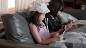 Two Asian girls are playing fun and exciting online mobile games at the gray sofa in their living room.