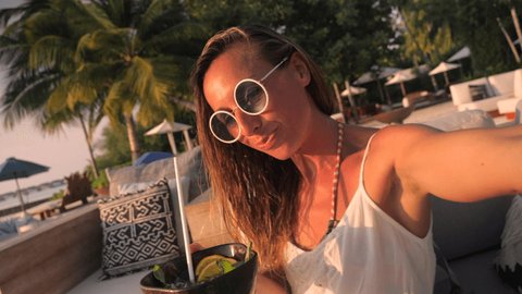 Young woman taking cool selfie at a beach bar wile having a tropical cocktail. Resort bar by the beach, female enjoying tropical vacation 