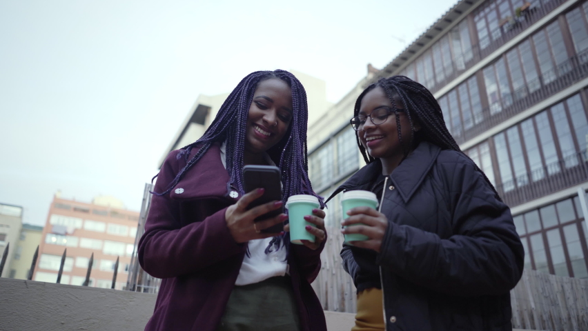 Two young women outside drinking coffee while discussing something they are watching on their cell phones. They laugh and chat during a break in their office. Royalty-Free Stock Footage #1068298487
