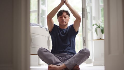 Mature Asian man in pyjamas sitting on bedroom floor stretching and meditating in yoga pose - shot in slow motion