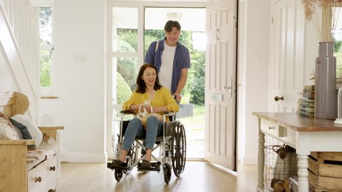 Mature Asian man pushing wife in wheelchair in hallway at home returning from shopping trip holding reusable bag - shot in slow motion