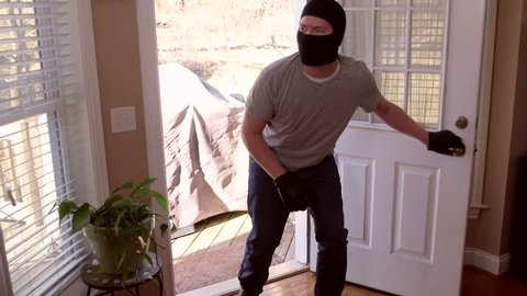A man who is a thief and burglar breaking into a house through the back door while wearing a ski mask and walking towards the camera to look directly into the lens close up and wide eye.