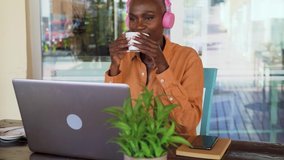Happy Afro senior woman having fun using computer and listening music with headphones while drinking coffee in bar restaurant - Digital nomad and freelance lifestyle concept