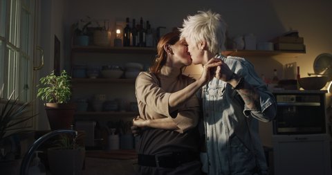 Cinematic shot of young carefree happy smiling homosexual female gay and transgender man couple in love is enjoying time together, dance together and kissing while cooking in kitchen at home.