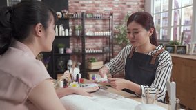 Summer manicure and nail color samples. Master woman manicurist helping customer chooses color of nail polish from palette with lady finger in nail beauty salon. smiling beautician listen client talk